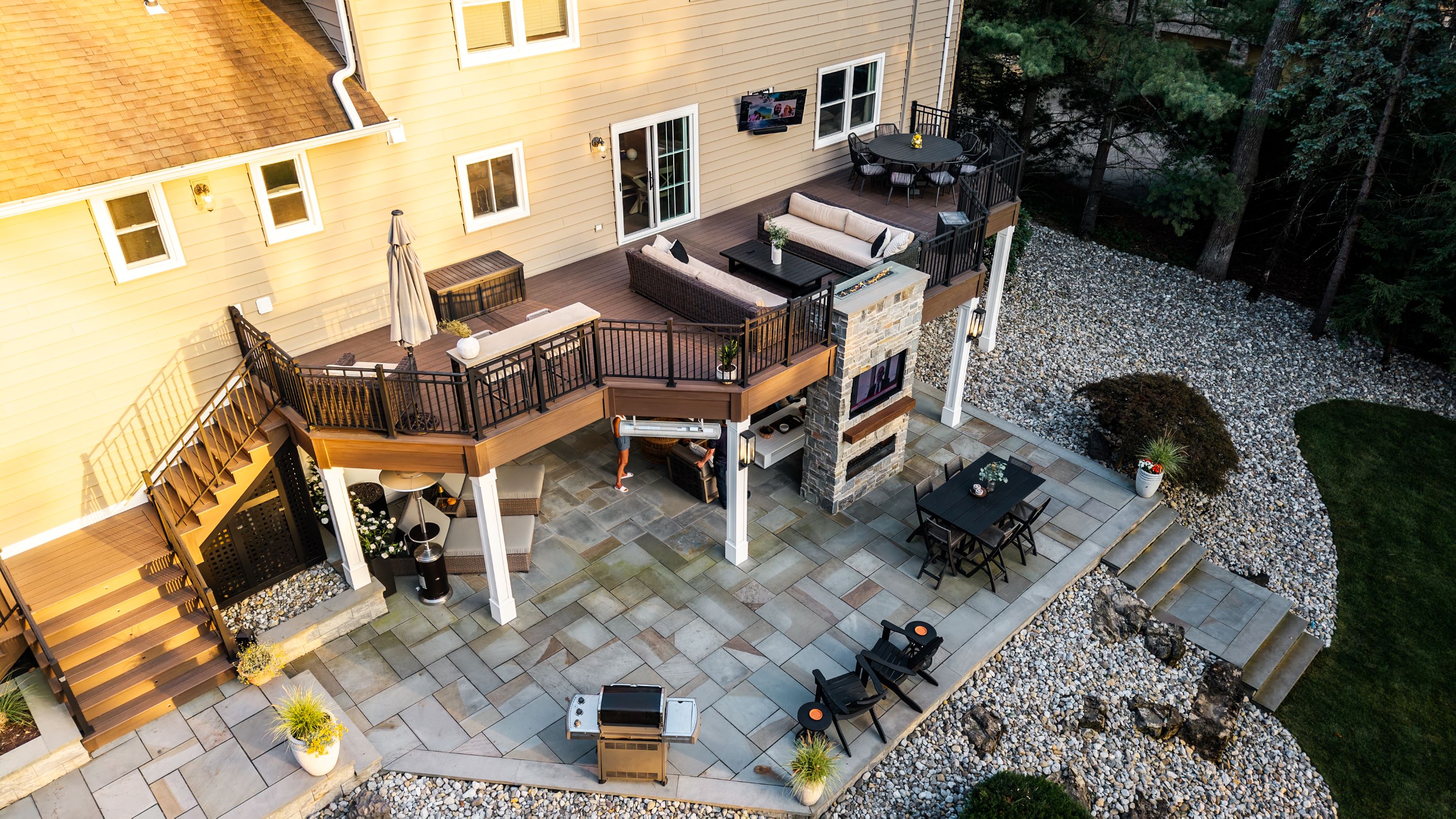 elevated deck with black railing and stairs above large paver patio featuring covered lounge area, outdoor fireplace, and dining space