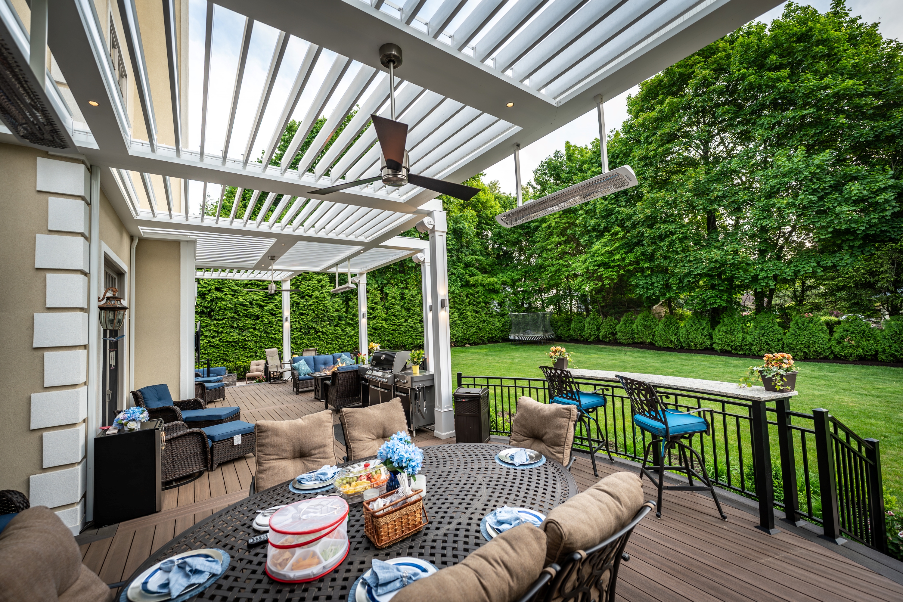 covered deck with louvered roof, dining table, grill station, lounge seating, and railing overlooking backyard lawn