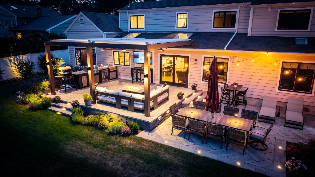 Backyard deck with covered lounge area, built-in grill, and dining patio lit by string lights at night, Made by Deck Remodelers